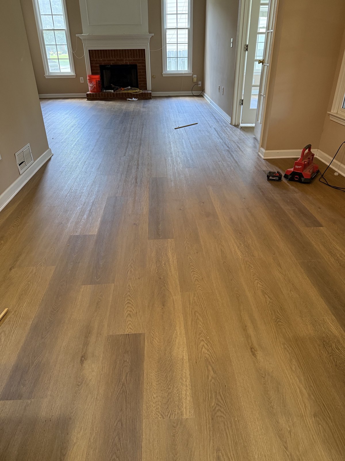 Empty living room with newly installed wood-look plank flooring, a brick fireplace, and a red power tool on the floor